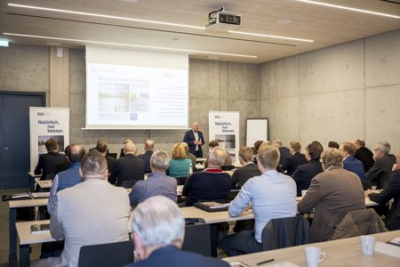 Ein Sprecher hält an einem Podium eine Präsentation vor einem Publikum in einem 