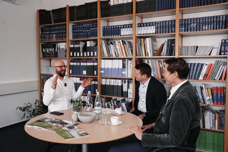 Drei Männer sitzen an einem Tisch in einem Büro mit Bücherregalen im Hintergrund